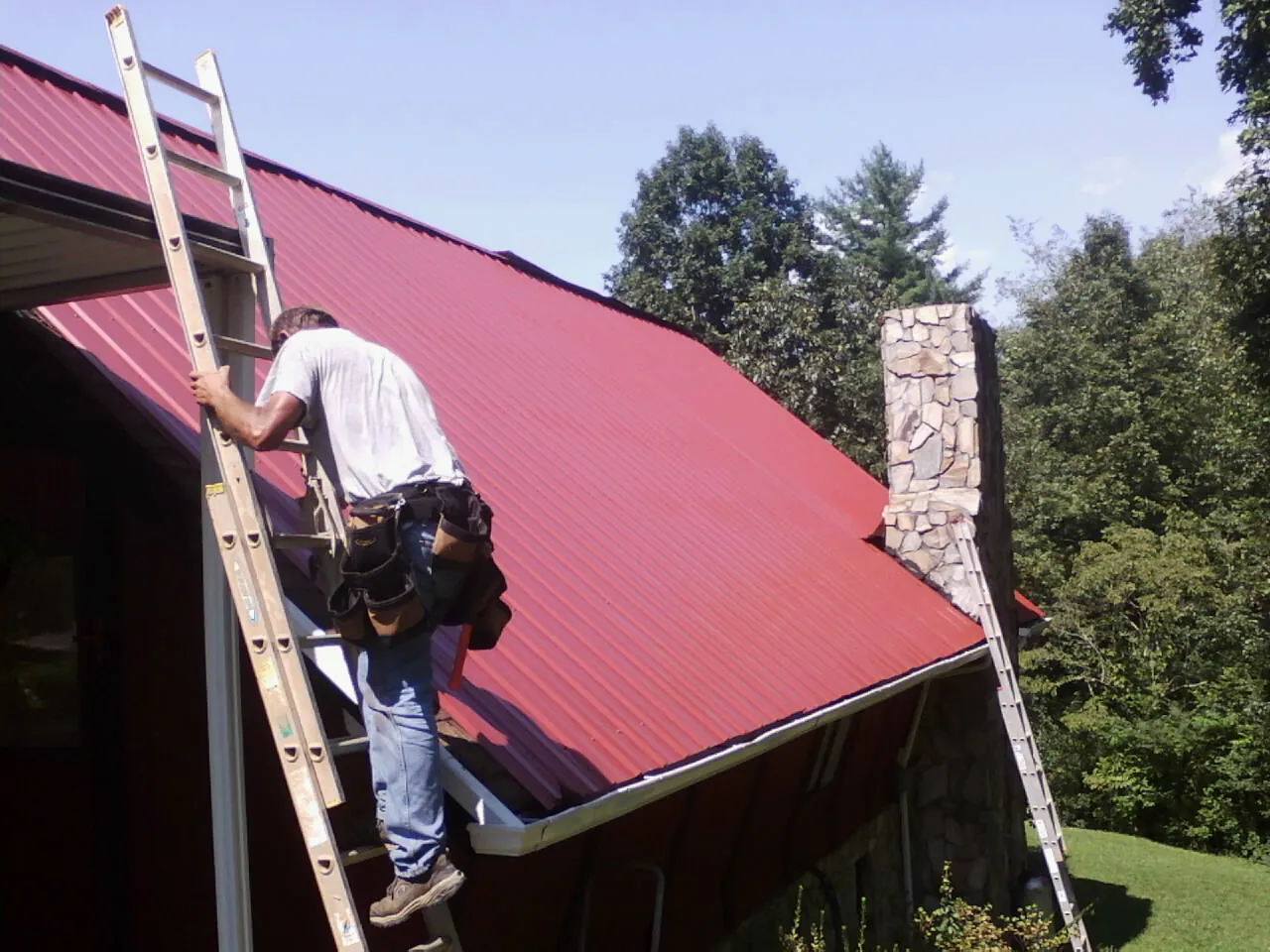 Roofer on a ladder beside a red metal roof and stone chimney — Soffit & Fascia Repair by Roofing Brothers in Azalea